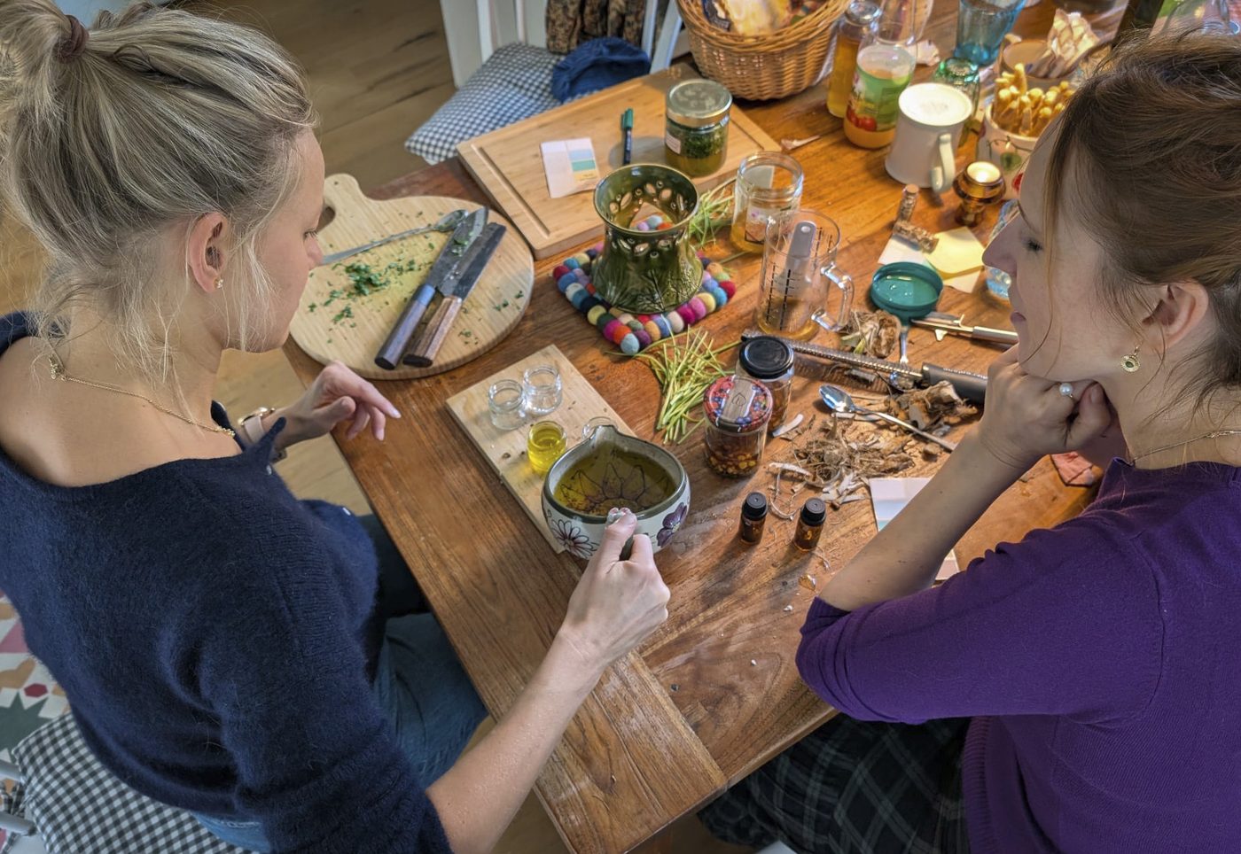 Zwei Frauen am Werktisch mit Kräutern, Gläsern und Ölen